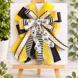May include: A decorative bow with a bee theme, displayed on a small wooden easel. The bow features yellow, black, and white ribbons with polka dots, stripes, and bee patterns. The bow is mounted on a white square.