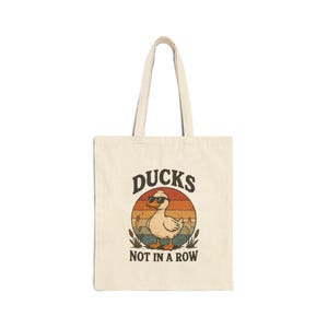 May include: A cream-coloured canvas tote bag featuring a graphic of a duck wearing sunglasses. The design includes a retro-style sunset background, cattails, and the text DUCKS NOT IN A ROW printed in a dark brown, bold, arched font above and below.