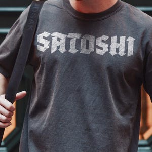 May include: Charcoal grey t-shirt featuring the word "SATOSHI" in a distressed silver font. The short-sleeved shirt is worn with a black shoulder bag, a wristwatch, and a beaded bracelet. The t-shirt appears to be made of a soft material.