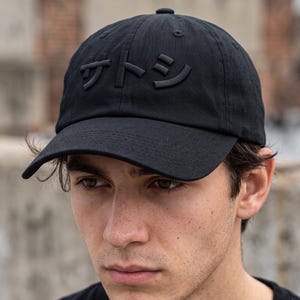 May include: A black baseball cap featuring the Japanese katakana characters for Satoshi embroidered in a matching black thread on the front panel. The cap has a structured crown, a curved brim, and a button at the top center of the headwear.