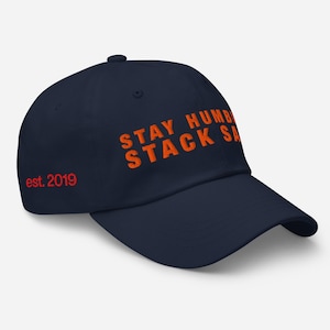 Puede incluir: Gorra de béisbol azul marino con el texto naranja "STAY HUMBLE STACK SA". El texto "est. 2019" está en rojo en el lateral. La gorra tiene una visera curva y un botón en la parte superior.