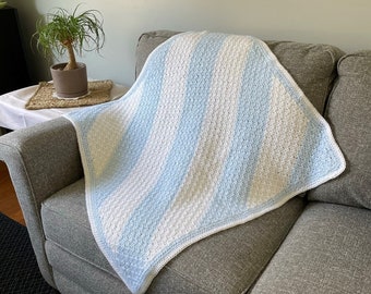 May include: A blue and white striped crocheted baby blanket draped over a gray couch.