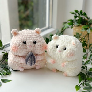 Puede incluir: Dos peluches de hámster tejidos a crochet, uno rosa y otro blanco, con ojos negros y mejillas rosadas. El hámster rosa sostiene una nuez tejida a crochet gris.