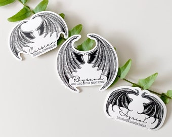 May include: Three black and white stickers with wings and names. The names are Cassian, Rhysand, and Azriel. The text below the names reads "General Commander", "High Lord of the Night Court", and "Spymaster Shadowsinger".
