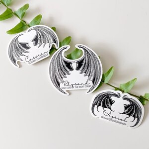 May include: Three black and white stickers with wings and names. The names are Cassian, Rhysand, and Azriel. The text below the names reads "General Commander", "High Lord of the Night Court", and "Spymaster Shadowsinger".
