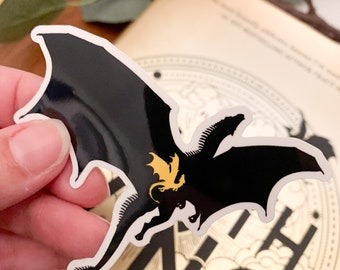 Op de afbeelding: Zwarte en gouden draken sticker met een witte omtrek. De draak vliegt met zijn vleugels uitgespreid. De sticker zit op een boek met het woord "thing" zichtbaar.