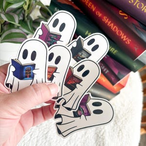 May include: Five white ghost stickers with black eyes, each holding a different book. The books are from the Throne of Glass series by Sarah J. Maas. The stickers are on a stack of books.