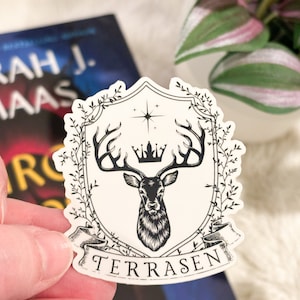 May include: A white sticker with a black and white illustration of a deer with antlers, a crown, and a banner that reads "Terrasen".
