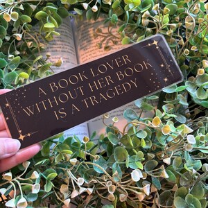 Może przedstawiać: Prostokątna czarna zakładka do książki ze złotym napisem A BOOK LOVER WITHOUT HER BOOK IS A TRAGEDY. Zakładka posiada złote akcenty w kształcie gwiazdek w rogach i jest umieszczona na tle zielonych liści eukaliptusa.
