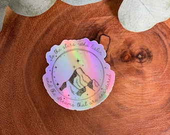 May include: A circular holographic sticker featuring a black line art illustration of mountain peaks surrounded by stars. The text reads, to the stars who listen and the dreams that are answered, written in a cursive font around the inner circle.
