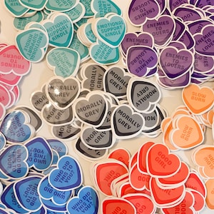 May include: Assortment of heart-shaped stickers with various sayings, including "Morally Grey", "Enemies to Lovers", "Slow Burn", and "Who Did This to You?" The stickers are in shades of blue, purple, gray, orange, and pink.