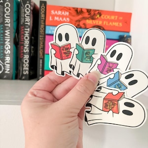 May include: Four white ghost stickers holding different colored books with the text "A Court of Thorns and Roses" on the covers. The stickers are on a white shelf with a hand holding them.