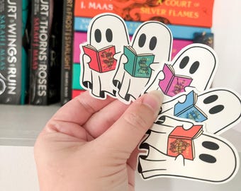 May include: Four white ghost stickers holding different colored books with the text "A Court of Thorns and Roses" on the covers. The stickers are on a white shelf with a hand holding them.