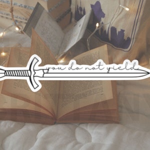 May include: A black and white sticker of a sword with the text "you do not yield" on a white background. The sticker is placed on an open book with a string of lights in the background.