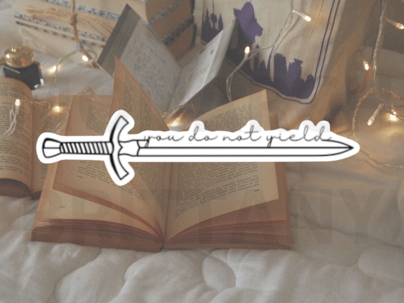 May include: A black and white sticker of a sword with the text "you do not yield" on a white background. The sticker is placed on an open book with a string of lights in the background.