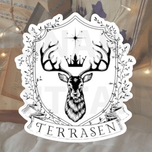 May include: Black and white illustration of a deer head with antlers inside a shield-shaped frame with a crown and the word "TERRASEN" at the bottom. The frame is surrounded by leafy branches.