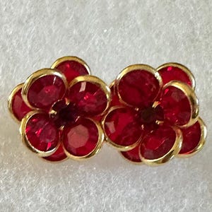 May include: A pair of flower-shaped earrings. Each earring features a cluster of red, faceted gemstones set in a gold-tone metal frame. The earrings are designed to resemble a flower.