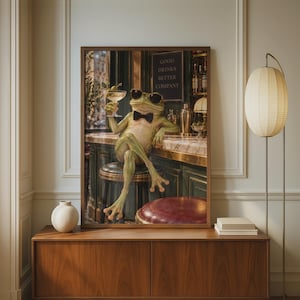 May include: A framed wall art print depicting a green frog wearing black sunglasses and a black bow tie, sitting on a bar stool while holding a martini glass. A sign in the background reads GOOD DRINKS BETTER COMPANY in a dimly lit bar setting.