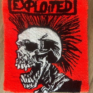 Op de afbeelding: Een vierkant, oranje tapijt met een zwart-wit schedelontwerp. Het woord "EXPLOITED" staat in een zwarte rechthoek bovenaan. De schedel heeft een hanenkam en kijkt naar links. Het tapijt heeft een getextureerd uiterlijk.