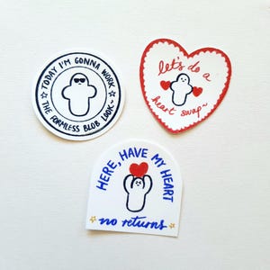Può includere: Tre adesivi bianchi con testo nero e rosso. Un adesivo circolare recita "Today I'm gonna work the formless blob look". Un adesivo a forma di cuore dice "Let's do a heart swap". Un terzo adesivo dice "Here, have my heart, no returns."