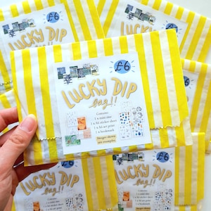 May include: A collection of yellow and white striped paper bags, each marked "Lucky Dip bag!!". The bags feature a sticker detailing contents: a mini zine, sticker sheet, art print, and bookmark. One bag is held in a hand.