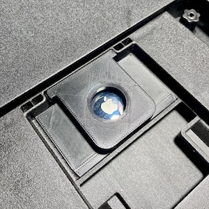 以下が含まれることがあります： 黒色のテクスチャード加工されたプラスチック部品のクローズアップ。円形の開口部があり、その中に白いリンゴのロゴが入った反射面があります。この部品は、より大きなデバイスの一部です。