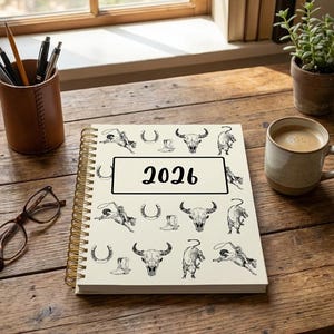 May include: A cream-coloured spiral-bound planner with black cowboy-themed illustrations and the year "2026" in a black rectangle. The planner sits on a wooden surface, alongside a coffee cup and a potted plant, near a window.