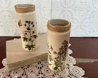 May include: Two cream-colored, cylindrical vases with floral and butterfly designs. The rims are wrapped with twine. One vase features a butterfly and dark floral print, the other has a purple floral design. The vases sit on a stack of paper and a lace doily.