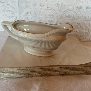May include: A white ceramic gravy boat with a gold trim and a decorative pattern. The boat is sitting on a stack of cardboard.