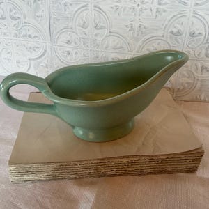 May include: A green ceramic gravy boat with a handle. The boat is sitting on a stack of brown cardboard.