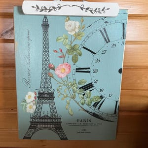 May include: A decorative clipboard featuring a vintage design with the Eiffel Tower, a clock face, and floral accents. The background is a light blue color. The clipboard has a white top with a floral design.