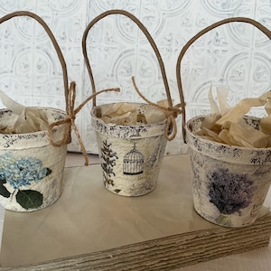May include: Three decorative, small, white buckets with brown handles and floral designs. Each bucket has a different floral image: blue hydrangea, a bird cage, and purple lavender. The buckets are filled with beige paper and tied with twine.