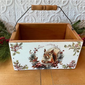 May include: A white wooden box with a wire handle and a painted design of a squirrel surrounded by holly berries and leaves. The box is perfect for storing small items or for use as a decorative piece.
