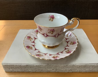 May include: A white teacup and saucer with a pink and burgundy floral pattern and gold trim. The cup has a delicate handle and the saucer has a scalloped edge.