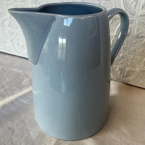 May include: A light blue ceramic jug with a smooth, glossy finish. The vessel features a wide, curved pouring spout and a sturdy, integrated handle on the side. The body of the jug is cylindrical with a slightly tapered base and a wide opening.