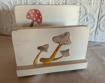 May include: White wooden magazine holder with a painted design of red and white mushrooms on the top and brown and white mushrooms on the bottom.