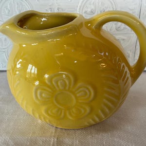 May include: A small, yellow ceramic pitcher with a rounded body and a curved handle. The pitcher features a floral design on the front and a spout for easy pouring. The pitcher is likely used for serving liquids.