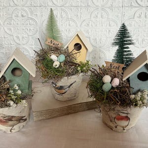 May include: Three decorative birdhouses with the word "Nest" on a sign, each in a pot with a bird design, surrounded by faux greenery and eggs.