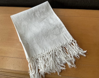 May include: A folded, off-white linen towel with a floral pattern and fringe trim. The towel is rectangular and features a repeating pattern of stylized flowers. The fringe is along one edge.