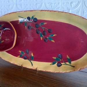 May include: An oval ceramic serving dish with a smaller round bowl attached. The dish is red with a yellow rim and olive branch designs in green, yellow, and black.