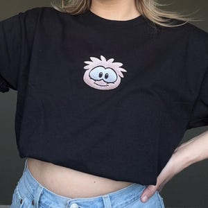 May include: A black short-sleeved t-shirt featuring a centered embroidered graphic of a light pink, round, spiky-haired character with large white eyes and a small smile. The shirt is worn with light blue denim jeans with a visible metal button.