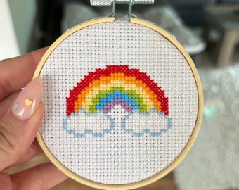 May include: A rainbow cross-stitch embroidery hoop with a white background and a rainbow with clouds.