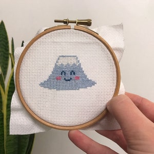 May include: A white embroidery hoop with a blue and white cross-stitch design of a smiling mountain with a white snowy peak.
