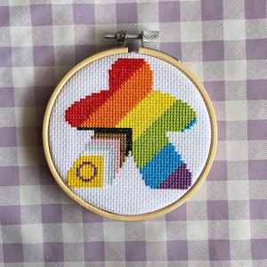 Op de afbeelding: Een kruissteekborduurwerk in een houten hoepel toont een kleurrijk spelstukontwerp. Het ontwerp bevat de kleuren van de regenboog en andere pride-vlaggen. De achtergrond is een witte stof met een paars-wit geblokt patroon.