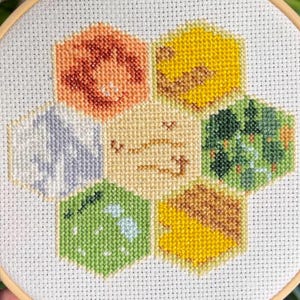 May include: A cross-stitch embroidery featuring a honeycomb design with seven hexagonal cells. The cells are filled with various colors, including orange, yellow, green, and gray, stitched on white fabric, and framed in a wooden hoop.