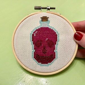 May include: Cross-stitch embroidery in a wooden hoop. The design features a skull inside a bottle, with a maroon skull and a light blue outline. The bottle has a cork stopper. The background is white.