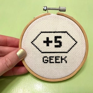May include: A white embroidery hoop with black thread, depicting a hexagonal shape with the text " +5 GEEK" inside.