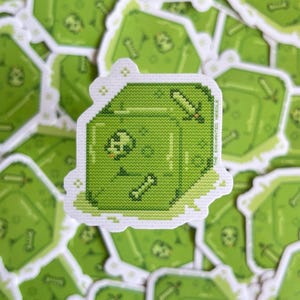 May include: A green pixelated sticker featuring a cube-shaped slime monster with a skull, bone, and sword. The sticker has a white border and is surrounded by other similar stickers. The overall theme is fantasy.