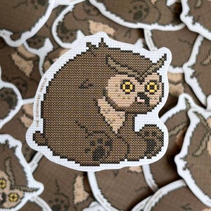 Può includere: Un adesivo con un gufo a punto croce nei toni del marrone e beige, occhi gialli e un bordo bianco. Il gufo è seduto. Il testo "THE ENCHANTED NEEDLE" è stampato sull'adesivo.