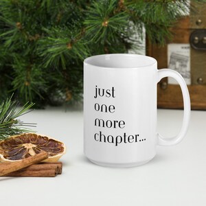 Könnte beinhalten: Weiße Keramik-Tasse mit dem Text "just one more chapter..." in Schwarz. Die Tasse hat einen gebogenen Henkel und steht vor einem Hintergrund mit Grün und einer Holzkiste.